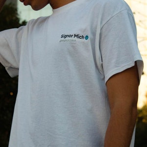 May include: White t-shirt with the text "Signor Mich" and a blue checkmark logo.