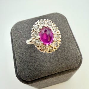 Pode incluir: Um anel de tom prateado com uma pedra central oval de cor rosa profundo em corte cabochão. A pedra central é rodeada por um halo de pequenas pedras transparentes de corte redondo, cravadas numa armação de metal com rebordo recortado.