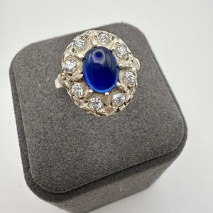 Pode incluir: Um anel de tom prateado com uma pedra central oval de cabochão azul profundo, rodeada por uma auréola de oito pequenas pedras transparentes de corte redondo, cravadas numa armação de metal texturizada e recortada.