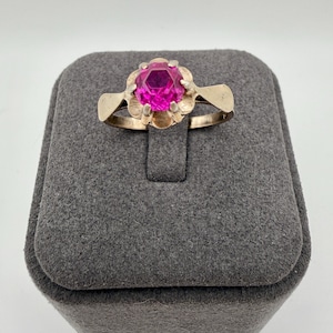 May include: A gold-toned ring featuring a large, faceted, magenta-colored gemstone. The gemstone is set in a floral-shaped setting. The ring is displayed on a gray, textured jewelry box.