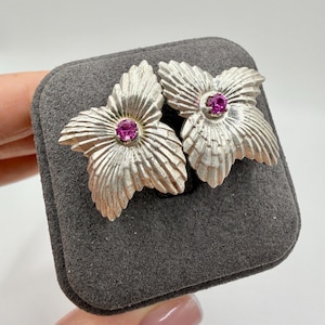 May include: A pair of silver-toned flower-shaped earrings with a textured finish. Each earring features a vibrant, round, magenta-colored gemstone at its center. The earrings are displayed on a gray, rectangular jewelry box.