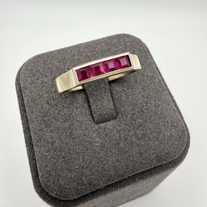 May include: A gold-toned ring featuring a rectangular design with four square, vibrant red gemstones. The ring is displayed on a dark gray jewelry box, showcasing its elegant design and color contrast.