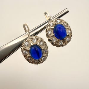 Pode incluir: Um par de brincos de prata com uma pedra azul oval em cabochão no centro, rodeada por um halo de pequenas pedras claras redondas facetadas. A estrutura de metal tem um acabamento texturizado e um fecho de alavanca articulado.