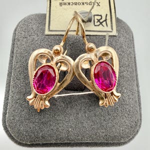 May include: A pair of gold-toned earrings with a heart-shaped design, each featuring a large, oval, magenta-colored gemstone. The earrings are displayed on a gray felt surface, with a tag visible in the background.