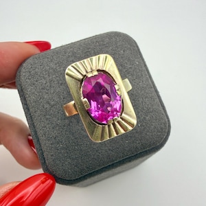 May include: A gold ring featuring a large, oval-cut, vibrant pink gemstone set in a rectangular gold plate with engraved sunburst-style lines radiating from the center stone. The ring band is plain gold and the setting is polished metal.