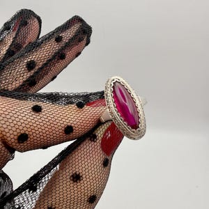 May include: A silver ring featuring a large, oval-shaped, magenta-colored stone. The stone is set in a decorative, openwork silver frame. The ring is displayed on a hand wearing a black, dotted lace glove.