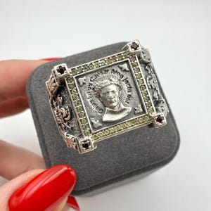 May include: A silver ring featuring a square face with a relief portrait of Julius Caesar and the inscription 'Gaius Julius Caesar Alea iacta est'. The border is set with small green stones, and each corner features a small dark red gemstone.