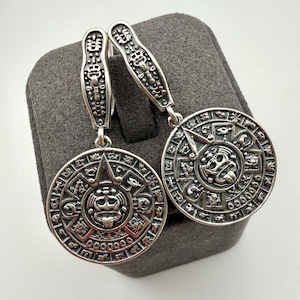 May include: A pair of silver drop earrings featuring circular pendants with intricate Aztec calendar motifs. Each earring has a long, textured silver hook with detailed engravings, connecting to a round medallion displaying a central face.