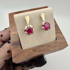 May include: A pair of gold-toned earrings with bright pink, round gemstones. Each earring features a floral-like setting around the gemstone and a simple, elongated top. The earrings are displayed on a beige surface within a wooden jewelry box.
