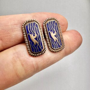 Op de afbeelding: Een paar gouden manchetknopen met een blauw emaille ontwerp. Het ontwerp toont een gestileerde vogel in vlucht tegen een achtergrond van golvende lijnen.