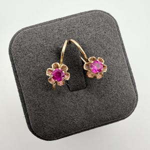 May include: A pair of gold earrings featuring a flower-shaped setting with a central round-cut vibrant pink gemstone. The earrings have curved wire hooks and are displayed on a dark gray square jewelry display pad with rounded corners.