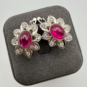 Pode incluir: Um par de brincos de prata com uma pedra central magenta em formato de cabochão oval, rodeada por uma moldura de inspiração floral cravejada com pequenas pedras redondas transparentes. Os brincos possuem um fecho de dobradiça seguro.