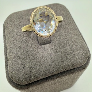 Pode incluir: Um anel com uma grande pedra preciosa transparente de corte oval e facetada, montada numa banda metálica. A pedra é fixada por um engaste metálico decorativo e recortado que rodeia o perímetro da gema, realçando a sua superfície multifacetada.
