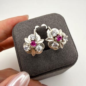 May include: A pair of silver-toned earrings featuring a floral-inspired design. Each earring is set with a central round-cut vibrant pink stone surrounded by three clear, colorless round-cut stones, all mounted on a polished silver-toned metal base.