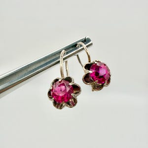 May include: A pair of silver-toned earrings featuring a flower design. Each earring has a bright pink, round gemstone set in a scalloped, dark-toned metal flower. The earrings are suspended from a simple hook.