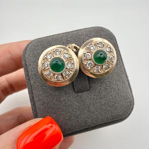 Pode incluir: Um par de brincos redondos de ouro com uma pedra cabochão verde central rodeada por um halo de pequenas pedras transparentes de corte redondo. Os brincos têm uma borda de ouro texturizada e estão num suporte quadrado de veludo cinzento.