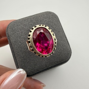 Può includere: Un anello color argento con una grande pietra preziosa dal taglio ovale di colore rosa fucsia vibrante. La montatura in metallo presenta un bordo testurizzato e dentellato attorno alla pietra centrale, che mostra molteplici sfaccettature.
