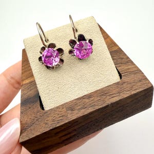May include: A pair of silver-toned leverback earrings featuring a central round-cut vibrant pink gemstone. Each stone is securely held within a metal setting shaped like a five-petaled flower, providing a floral aesthetic to the jewelry piece.