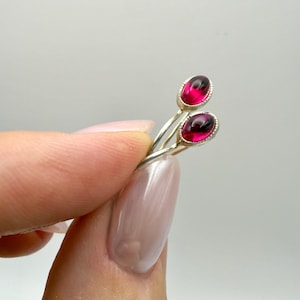 May include: A silver ring featuring two oval-shaped, deep pink cabochon gemstones set in textured metal bezels. The band is composed of two thin, polished silver wires that curve and overlap to hold the stones in a bypass-style arrangement.