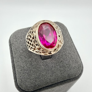 May include: A gold-toned ring with an oval, bright pink gemstone. The ring has an open filigree design around the gemstone. The ring is displayed on a gray, textured surface.