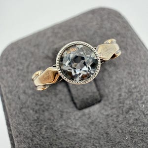 May include: A ring featuring a round, faceted, clear stone set in a silver-toned metal bezel with a milgrain edge. The band has a smooth, polished finish with a slightly twisted or contoured design that tapers towards the central setting.