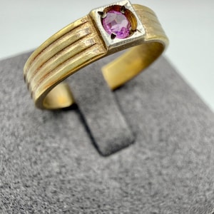 May include: A gold-toned ring with a square setting holding a round, pink gemstone. The band has horizontal ridges. The ring is displayed on a gray surface.