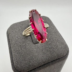 May include: A silver-toned ring featuring a large, rectangular, faceted, ruby-red gemstone. The ring has a classic design with a band that splits into two prongs on each side of the gemstone. The ring is displayed on a gray jewelry box.