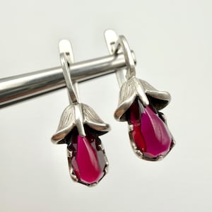 May include: A pair of silver-toned earrings featuring a floral design. Each earring has a flower-shaped top and a teardrop-shaped, deep magenta gemstone. The earrings are displayed against a white background.