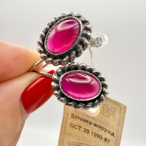 Puede incluir: Un par de anillos plateados con piedras preciosas ovaladas de color magenta. Las piedras están engastadas en un borde texturizado en forma de cuerda. Se adjunta una etiqueta con texto cirílico.
