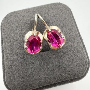 May include: A pair of silver-toned earrings featuring oval-cut, vibrant pink faceted stones. Each stone is securely held in a prong setting with a curved metal hook for attachment. The earrings have a polished metallic finish and a classic design.