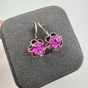May include: A pair of gold-toned earrings with a flower design. Each earring features a large, round, bright pink gemstone at the center, surrounded by smaller, darker pink petals. The earrings are displayed on a gray, textured surface.