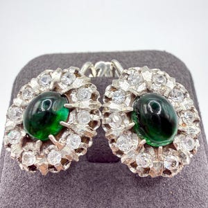 Pode incluir: Um par de brincos ovais com uma pedra cabochão verde-escura central rodeada por um halo de pequenas pedras brancas transparentes e facetadas, montadas numa estrutura de metal prateado texturado com um mecanismo de fecho de alavanca.