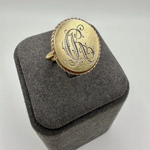 Puede incluir: Un anillo de sello dorado con una cara grande y circular. La cara presenta una letra "R" grabada en una fuente de escritura. El anillo tiene un borde en forma de cuerda y se muestra en una caja de joyería gris.