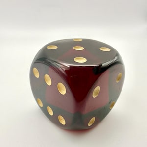 May include: A large, oversized die with a glossy, dark red finish. The die features gold-colored dots, with the number of dots varying on each face. The die is a decorative item, likely for display or novelty purposes.