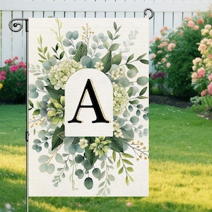 May include: A garden flag displaying the letter "A" in black, framed by a white arched shape. The flag is decorated with a floral design of green and white leaves and flowers, offering a natural, decorative aesthetic.
