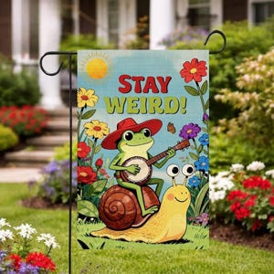 Puede incluir: Una bandera de jardín con una ilustración caprichosa. Una rana verde con un sombrero rojo toca el banjo sobre un caracol. La bandera dice "STAY WEIRD!" y está decorada con flores coloridas y un sol.
