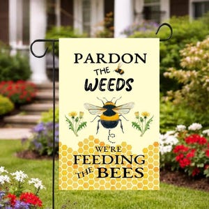 Op de afbeelding: Een gele tuinvaandel met de tekst "Pardon the Weeds, We're Feeding the Bees." De vlag toont een grote hommelillustratie, paardenbloemen en een honingraatpatroon. De vlag hangt aan een zwarte metalen standaard in een tuin.