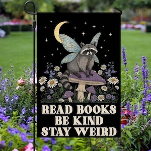 May include: A black garden flag featuring a raccoon with fairy wings sitting on a mushroom under a crescent moon and stars. The flag has the text "READ BOOKS BE KIND STAY WEIRD" in white letters. Daisies and other flowers surround the raccoon.
