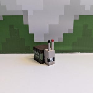 May include: A Minecraft-style toy snail figurine. The snail has a grey head with black and white pixelated details, a brown body, and two vertical antennae, one black and one red. The background features a pixelated green and white design.