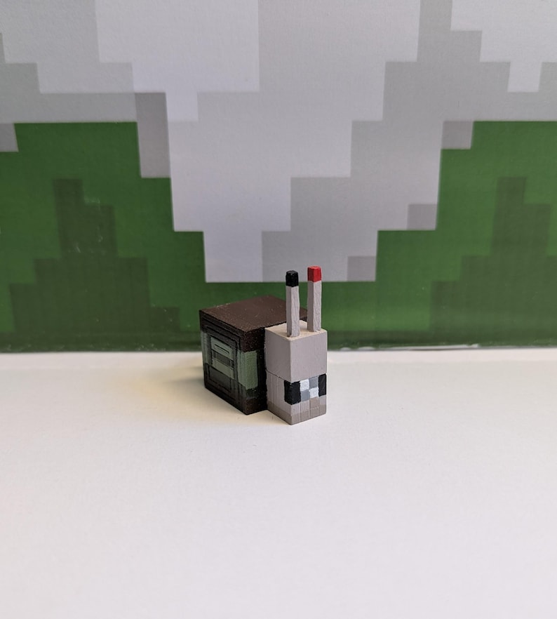 May include: A Minecraft-style toy snail figurine. The snail has a grey head with black and white pixelated details, a brown body, and two vertical antennae, one black and one red. The background features a pixelated green and white design.
