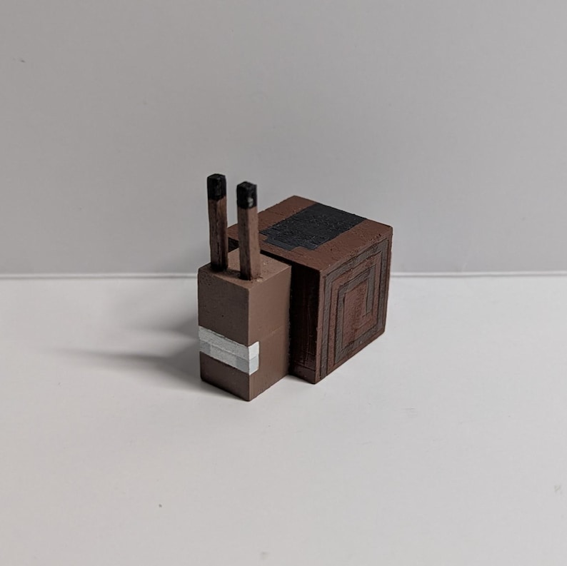 May include: A brown, blocky snail figurine with a square body and a rectangular head. The snail has two black antennae and a white stripe on its head. The body features a series of concentric square patterns.