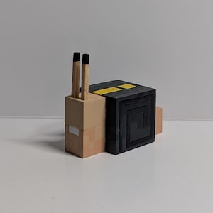 May include: A wooden snail figurine with a tan body and a black shell. Two matchsticks act as the snail's antennae. The shell has a square design with yellow stripes on top. The figurine is on a white surface.
