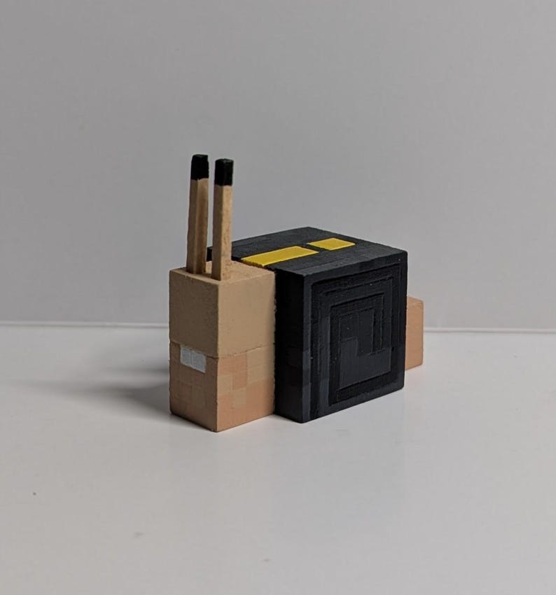 May include: A wooden snail figurine with a tan body and a black shell. Two matchsticks act as the snail's antennae. The shell has a square design with yellow stripes on top. The figurine is on a white surface.