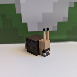 May include: A Minecraft-style snail figurine. The snail has a brown shell, a tan body, and two black-tipped antennae. The snail's face is a pixelated design with black, white, and brown squares. The background features a green and white pixelated design.