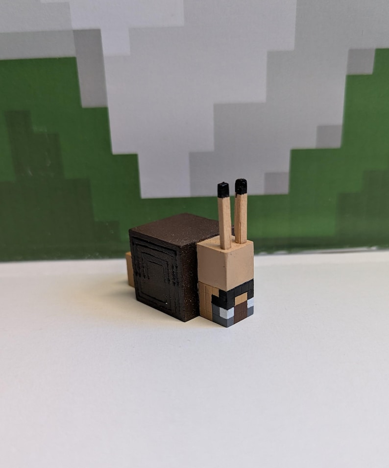 May include: A Minecraft-style snail figurine. The snail has a brown shell, a tan body, and two black-tipped antennae. The snail's face is a pixelated design with black, white, and brown squares. The background features a green and white pixelated design.