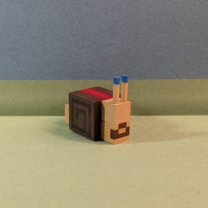 May include: A pixelated wooden snail ornament. The snail has a tan body and a dark brown shell with a red stripe. The snail's antennae are tan with blue tips. The snail is a handmade craft.