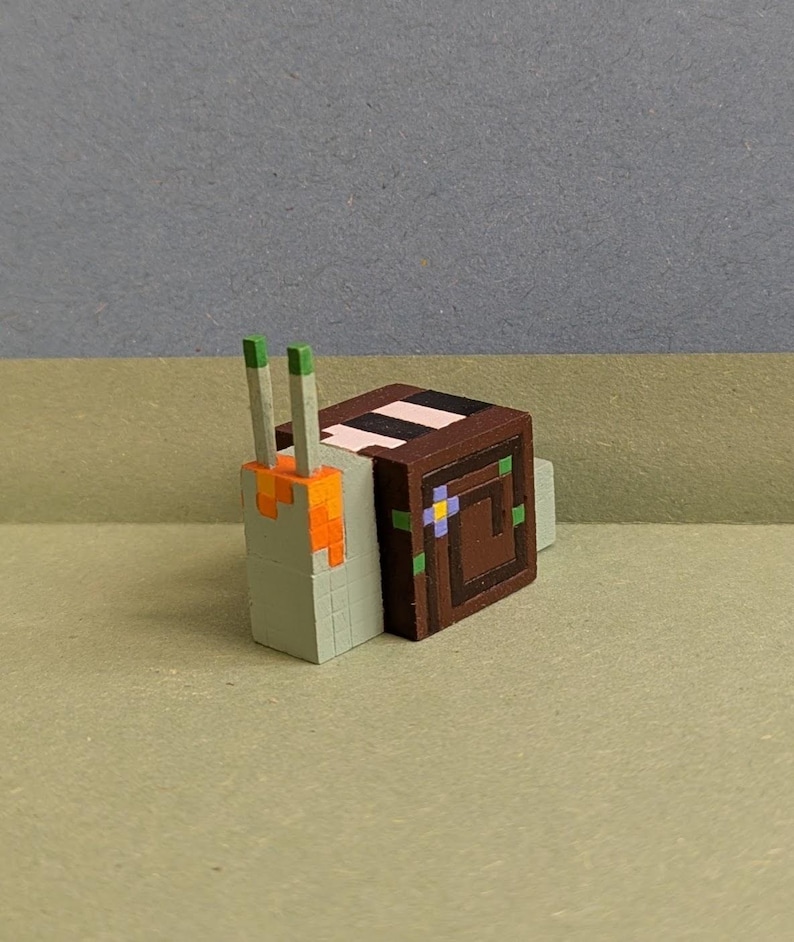 May include: A pixelated snail figurine with a brown shell and a light green body. The shell has a decorative pattern with a pink stripe and a square design. The snail's antennae are green and orange.