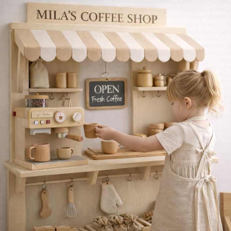 May include: Wooden toy coffee shop with a striped awning and the text "MILA'S COFFEE SHOP". Includes a toy espresso machine, cups, and a chalkboard sign that says "OPEN Fresh Coffee". A child is holding a wooden cup.