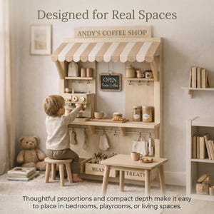 May include: A wooden play coffee shop with a striped awning, labeled "ANDY'S COFFEE SHOP." It includes a small table, stool, and accessories. The sign reads "OPEN Fresh Coffee." A child is interacting with the coffee maker. The text says "Designed for Real Spaces."