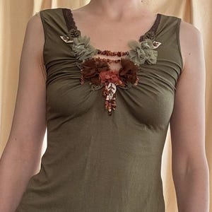 May include: Olive green tank top with decorative floral embellishments around the neckline. The top features a scoop neck and a fitted silhouette. The floral details include brown, green, and red accents, adding a touch of elegance to the garment.